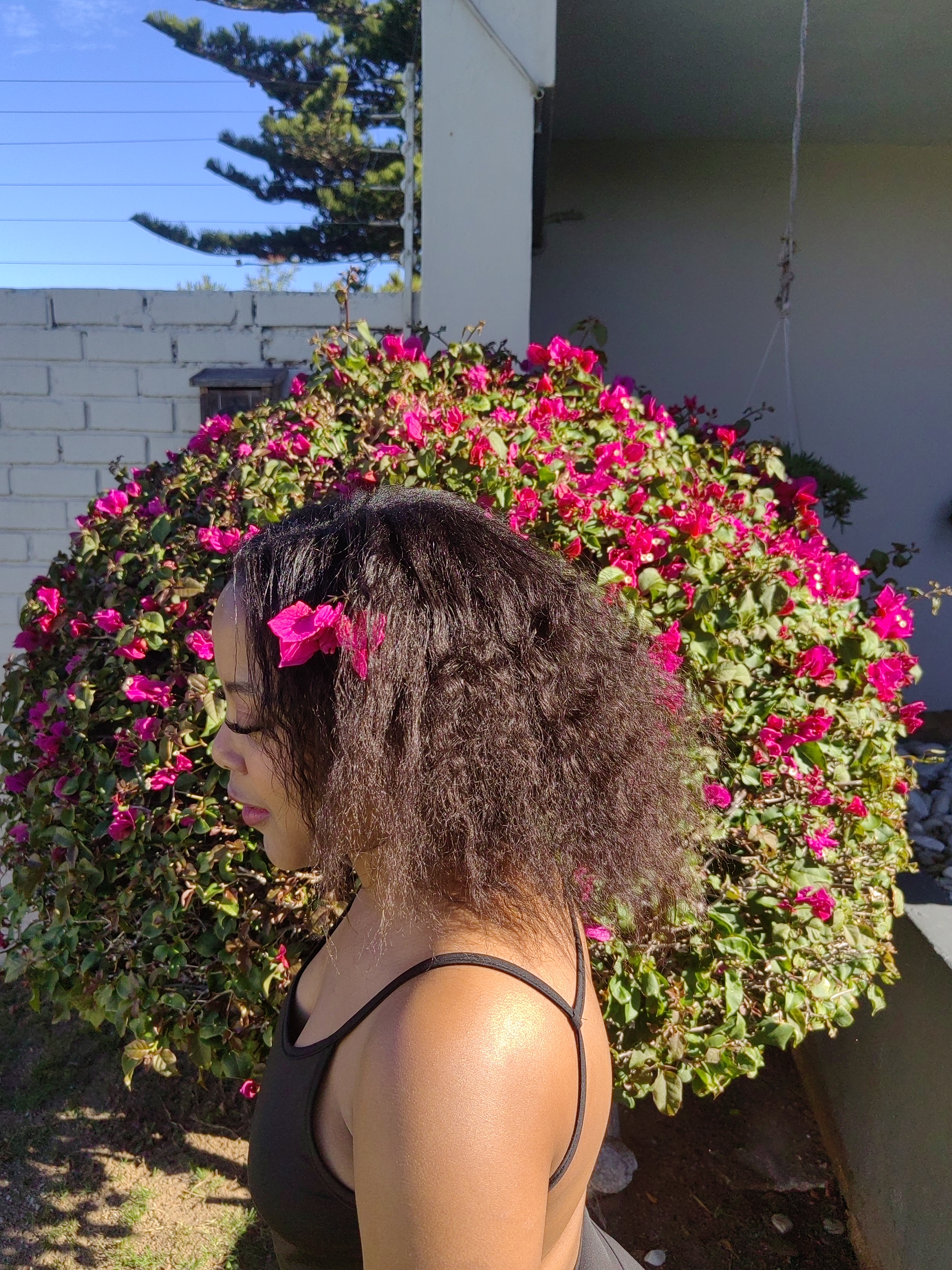 Nanda in profile, natural hair with a bougainvillea bloom tucked in · the hair journey that became Sanyu.